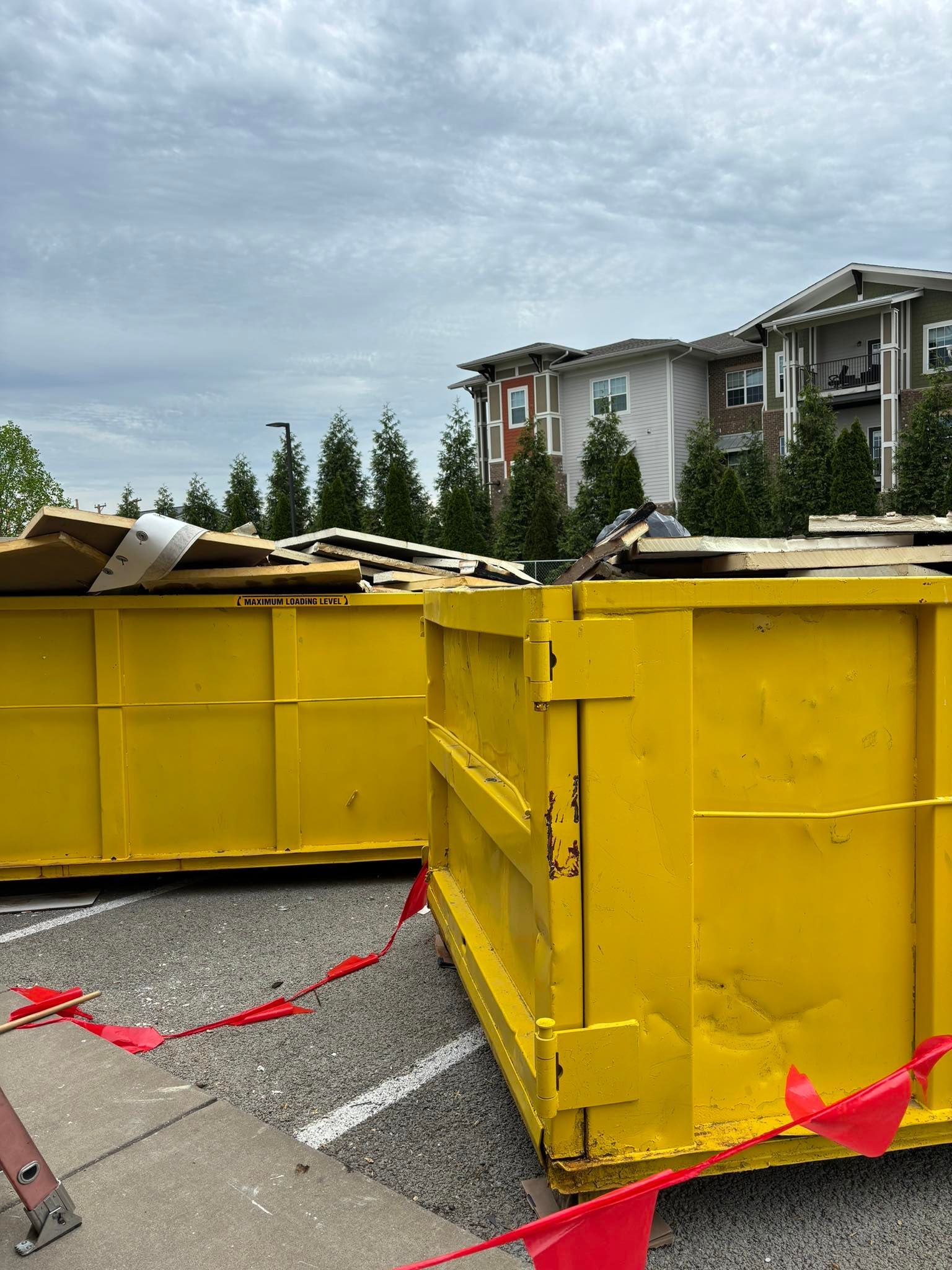 Two yellow dumpsters filled with debris sit in a parking lot. Apartment building in the background under a cloudy sky.