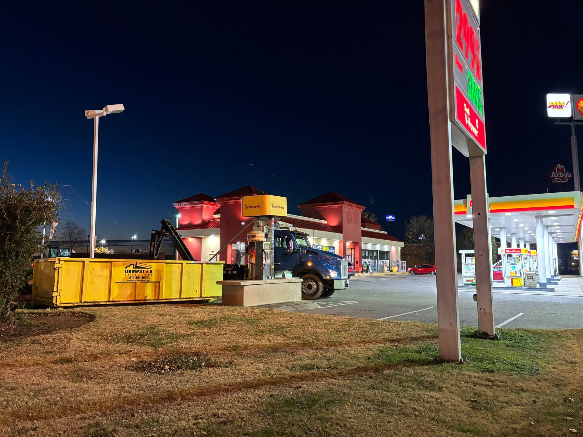 A dumpster truck beside a yellow dumpster at night in front of a Shell gas station and a red-roofed restaurant.