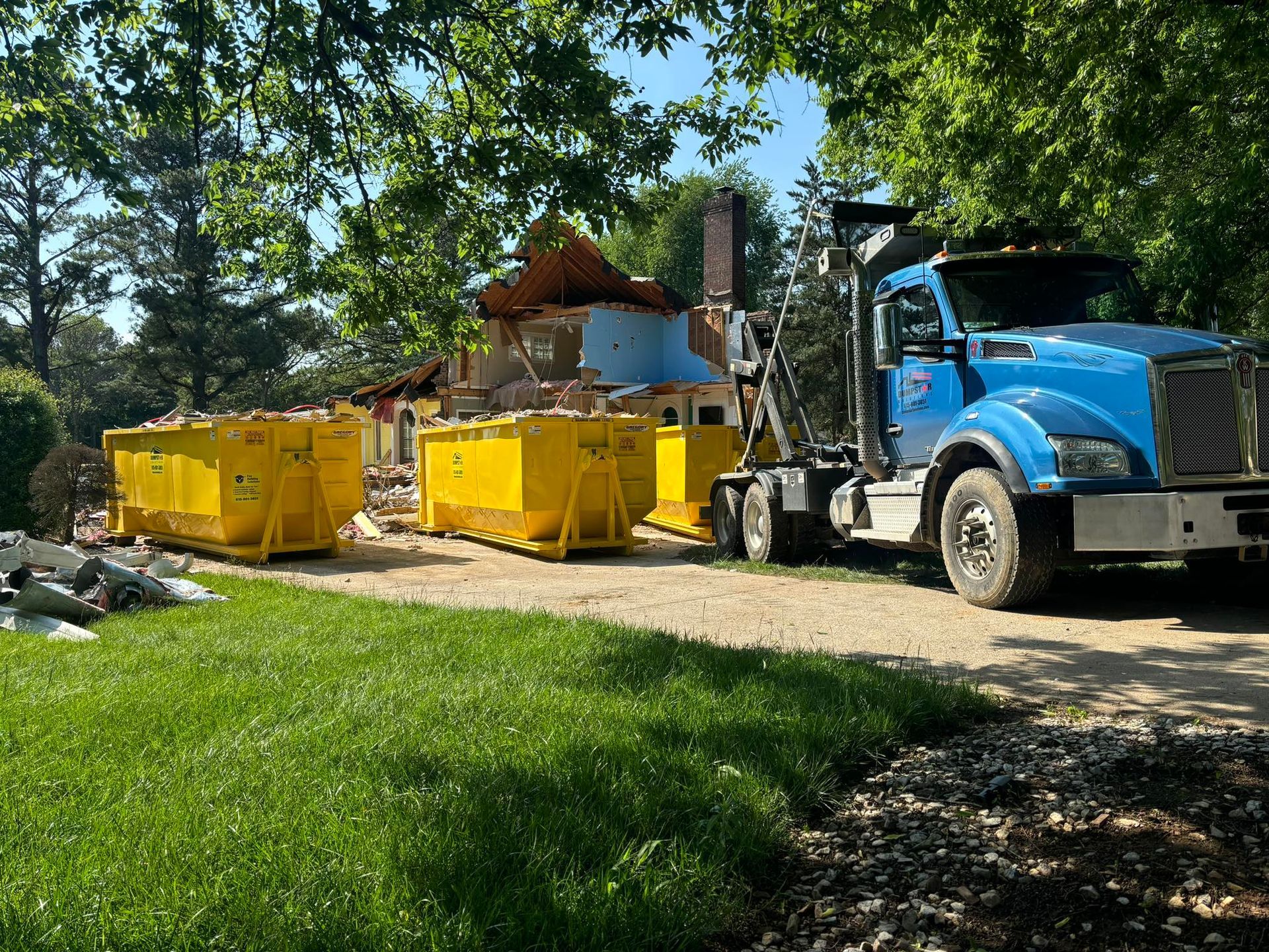 A blue truck next to yellow dumpsters at a house demolition site; debris and green grass.