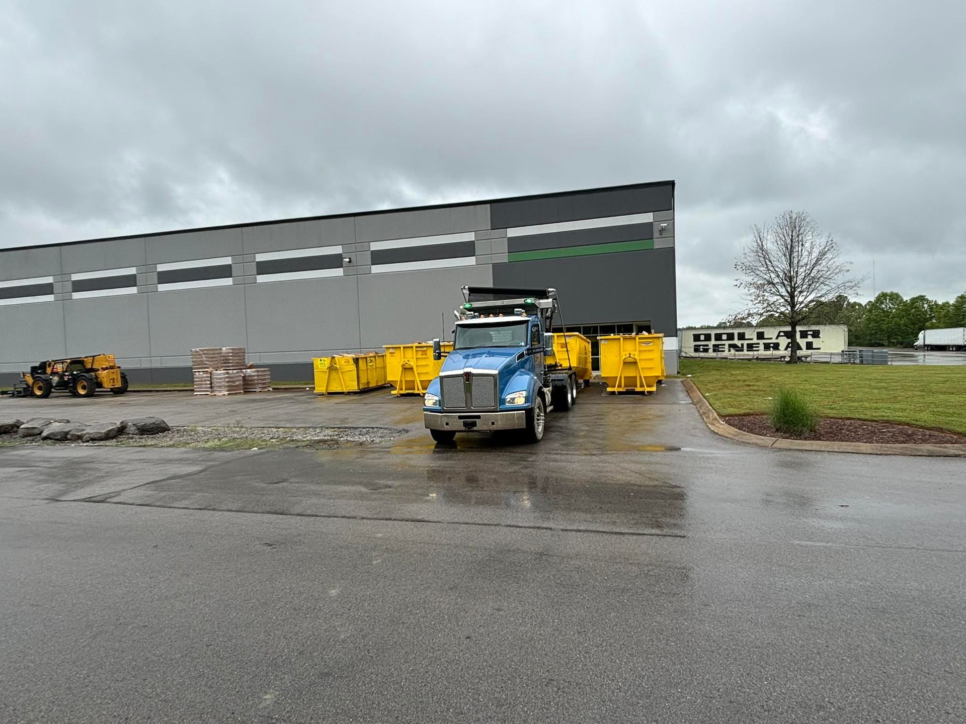 A blue truck with yellow bins parked in front of a large building on a cloudy day.