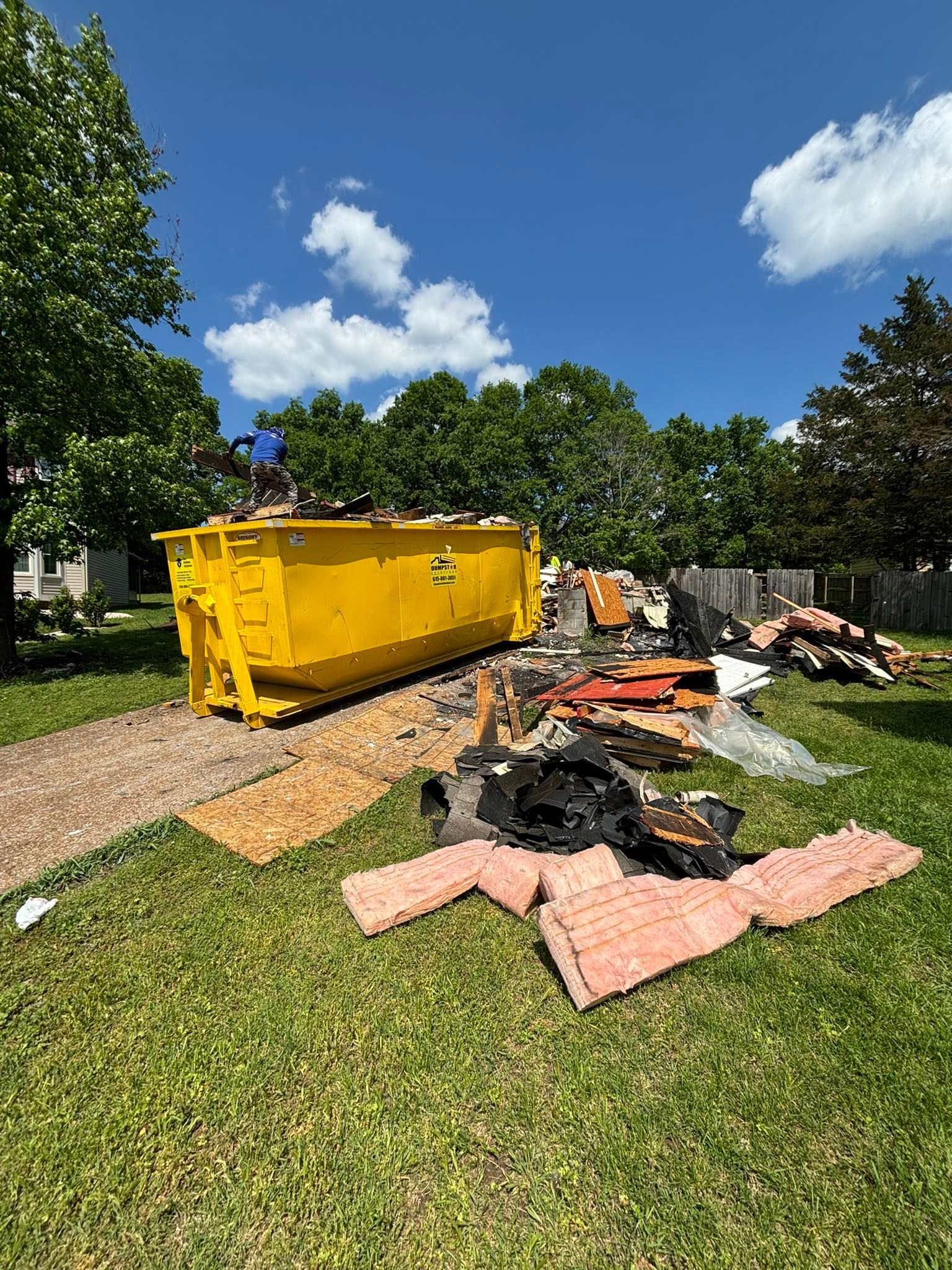 Yellow dumpster with debris on green grass; person loading it on a sunny day.