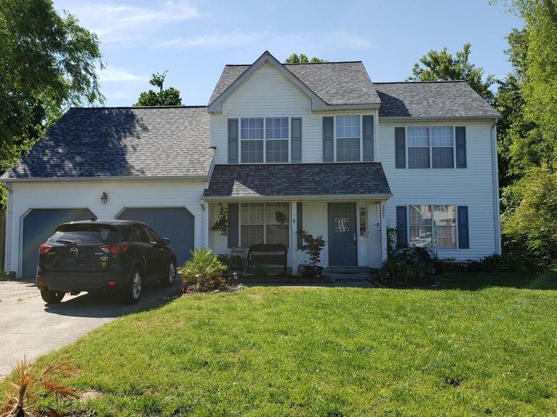 Two-story white house with blue shutters, a dark SUV in driveway, green lawn, blue sky.