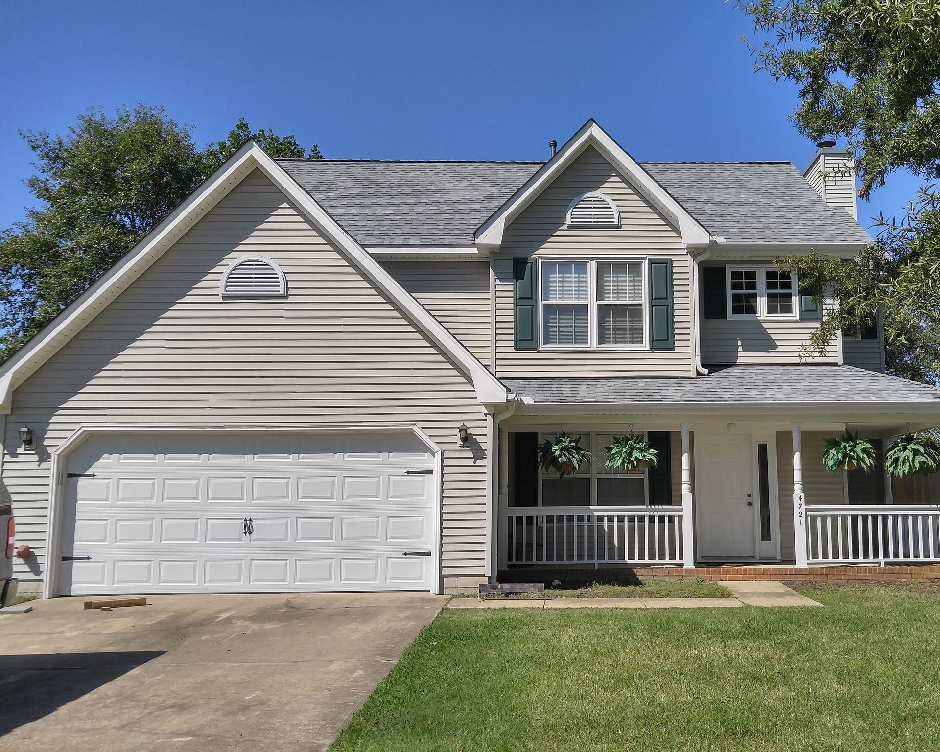 A two-story, light-colored suburban house with a two-car garage, a front porch, and a grassy yard under a clear blue sky.