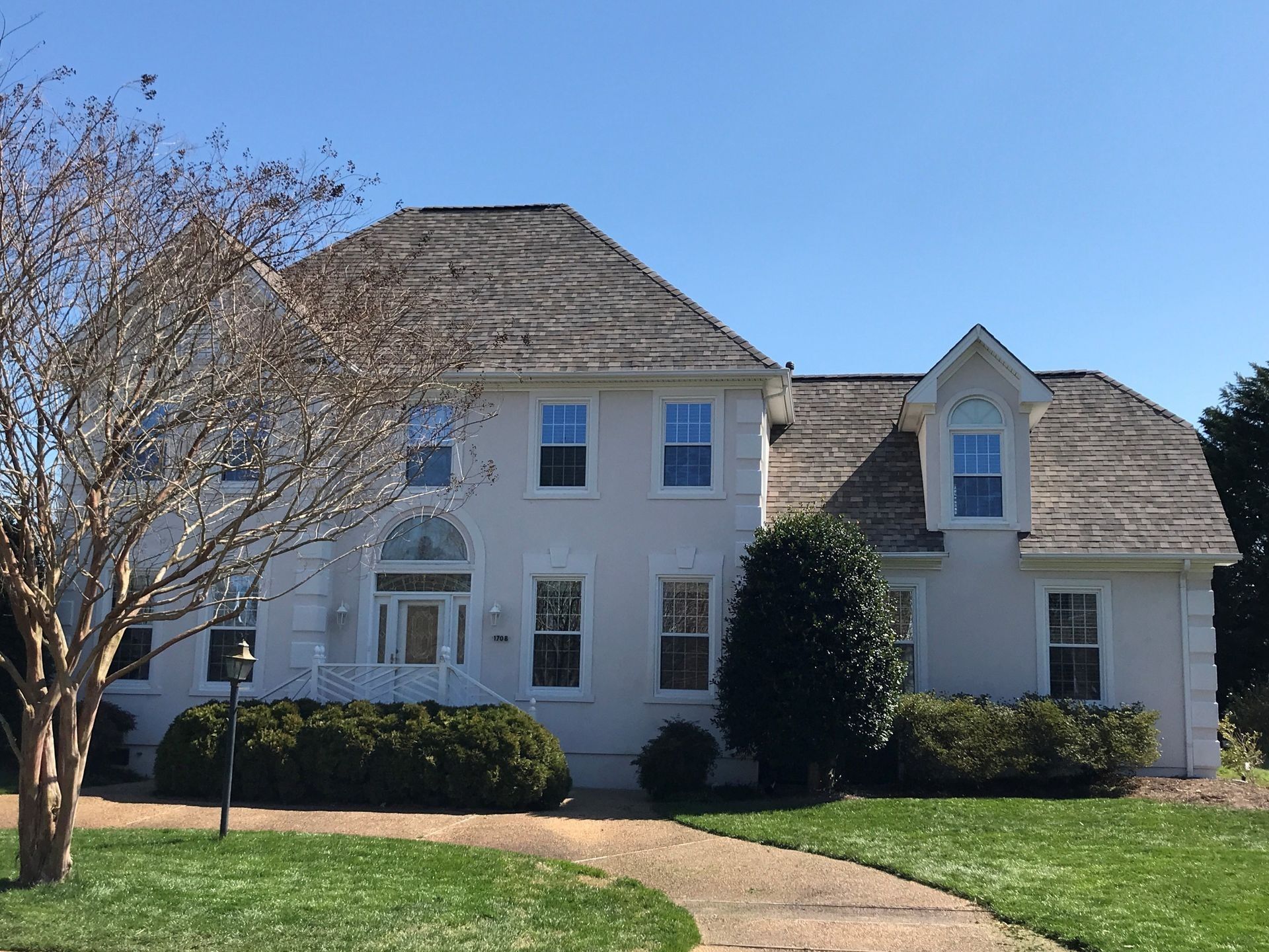 A two-story white house with a gray shingled roof, arched entryway, and dormer window, surrounded by a lawn and shrubs.