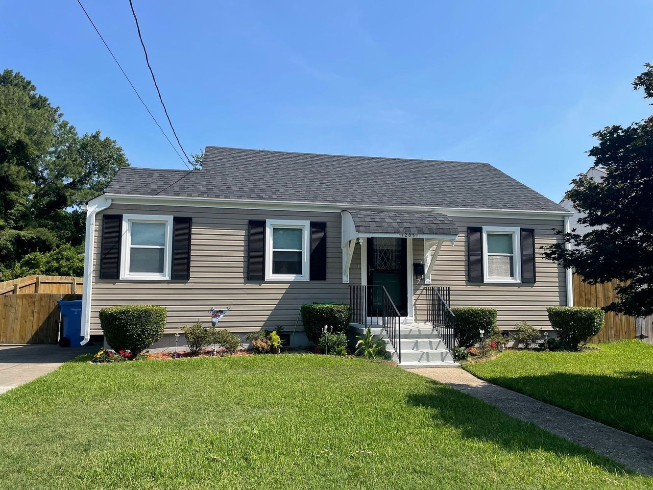 Tan-sided house with black shutters, gray roof, and green lawn under a blue sky.