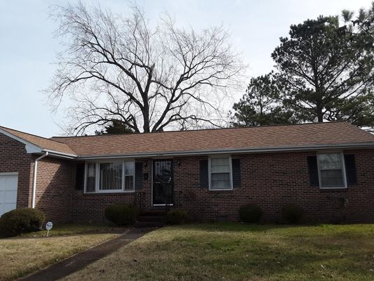 Brick house with brown roof, black shutters, and bare tree in background.