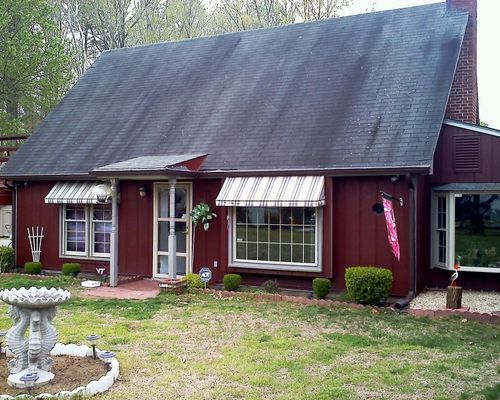 Red house with dark roof and awnings; a brick chimney, a bay window, and a lawn with a bird bath are visible.
