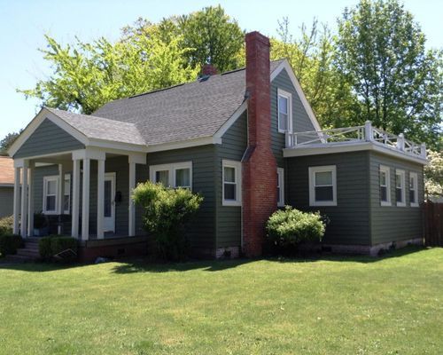 Green house with white trim, red brick chimney, and porch. Located on a grassy lawn with trees in the background.