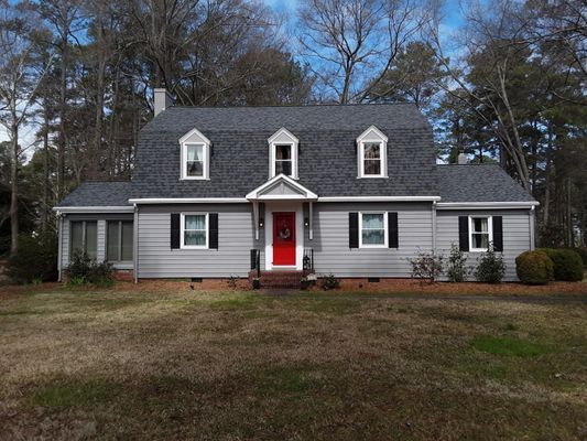 Gray house with red door, black shutters, and dormer windows on a cloudy day.