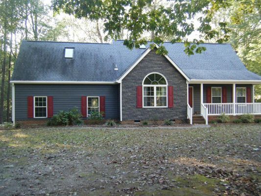 Gray house with red shutters, white trim, and a small porch, set in a wooded area.