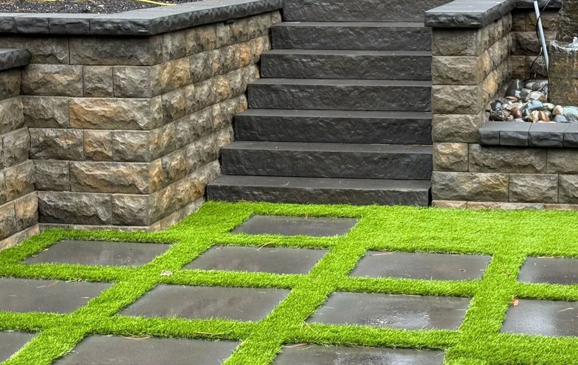 Stone steps and retaining walls next to a patio with square grass and paving stone pattern.