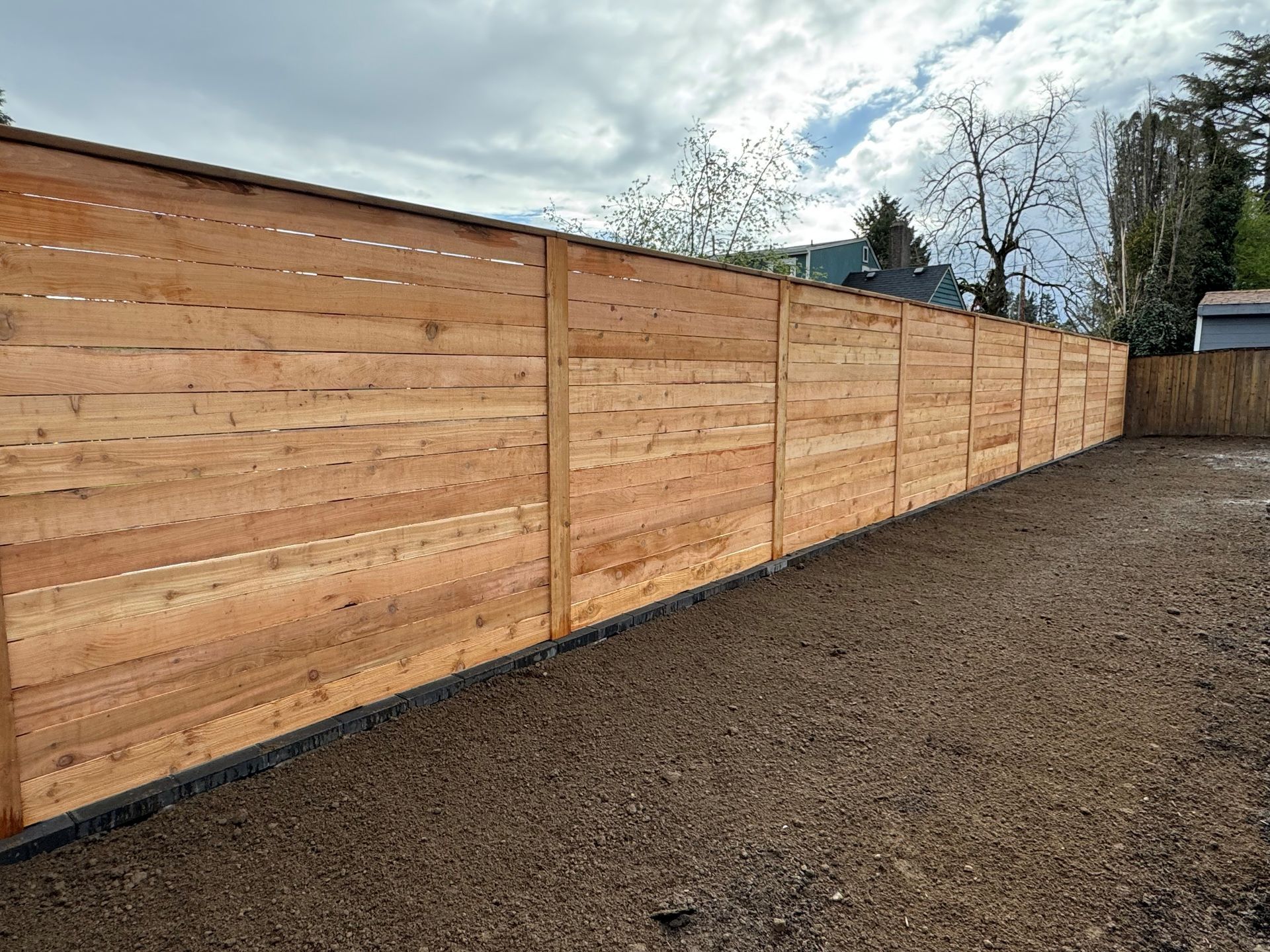 Wooden fence enclosing a yard with gravel ground and a cloudy sky in the background.