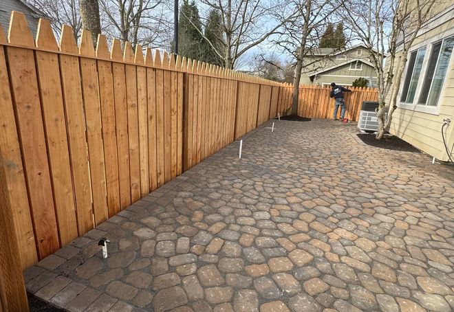 Stone patio next to a wooden fence and house. A person works in the background.