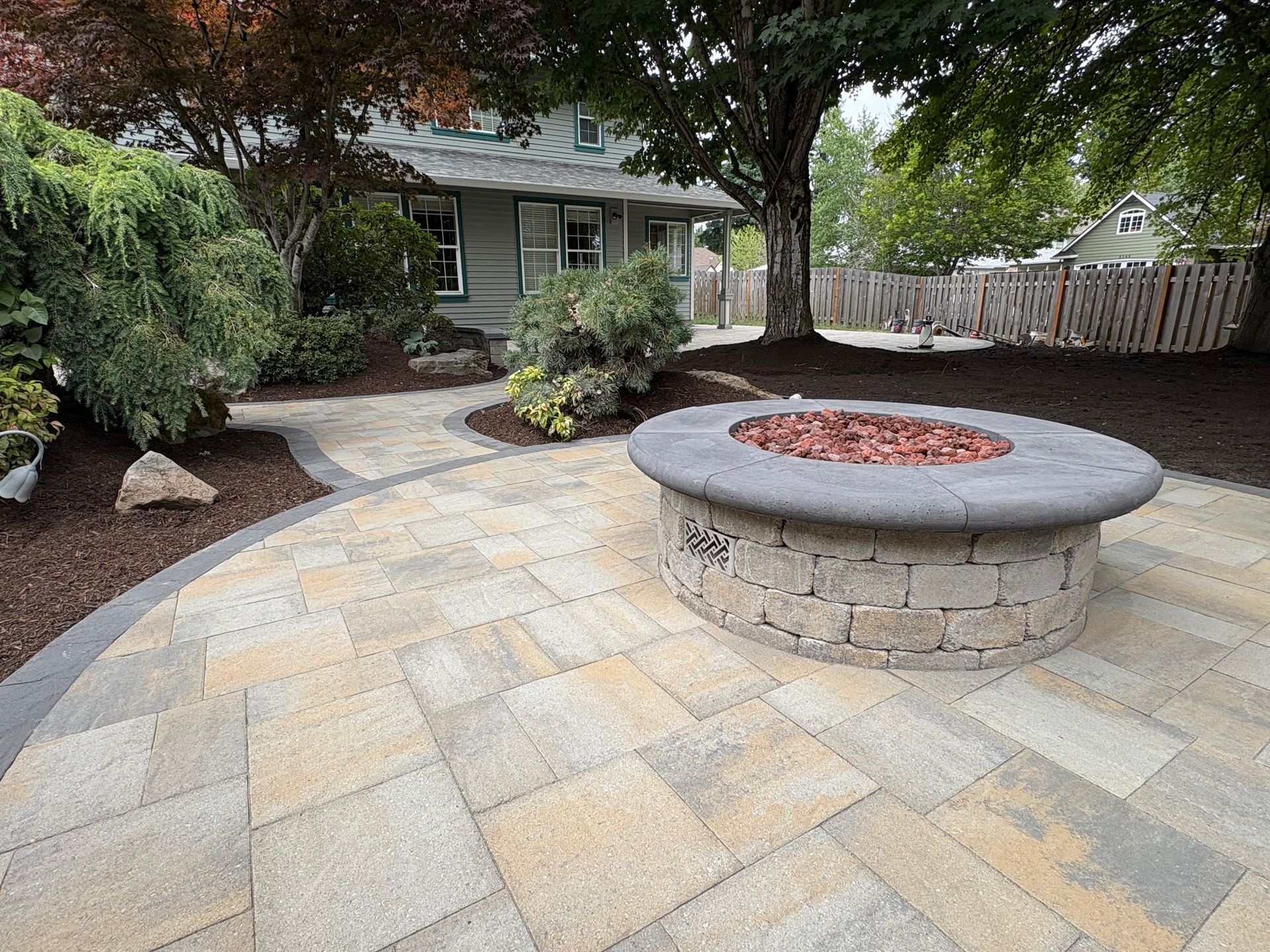 Stone patio with fire pit, pathway, and landscaping in front of a house.