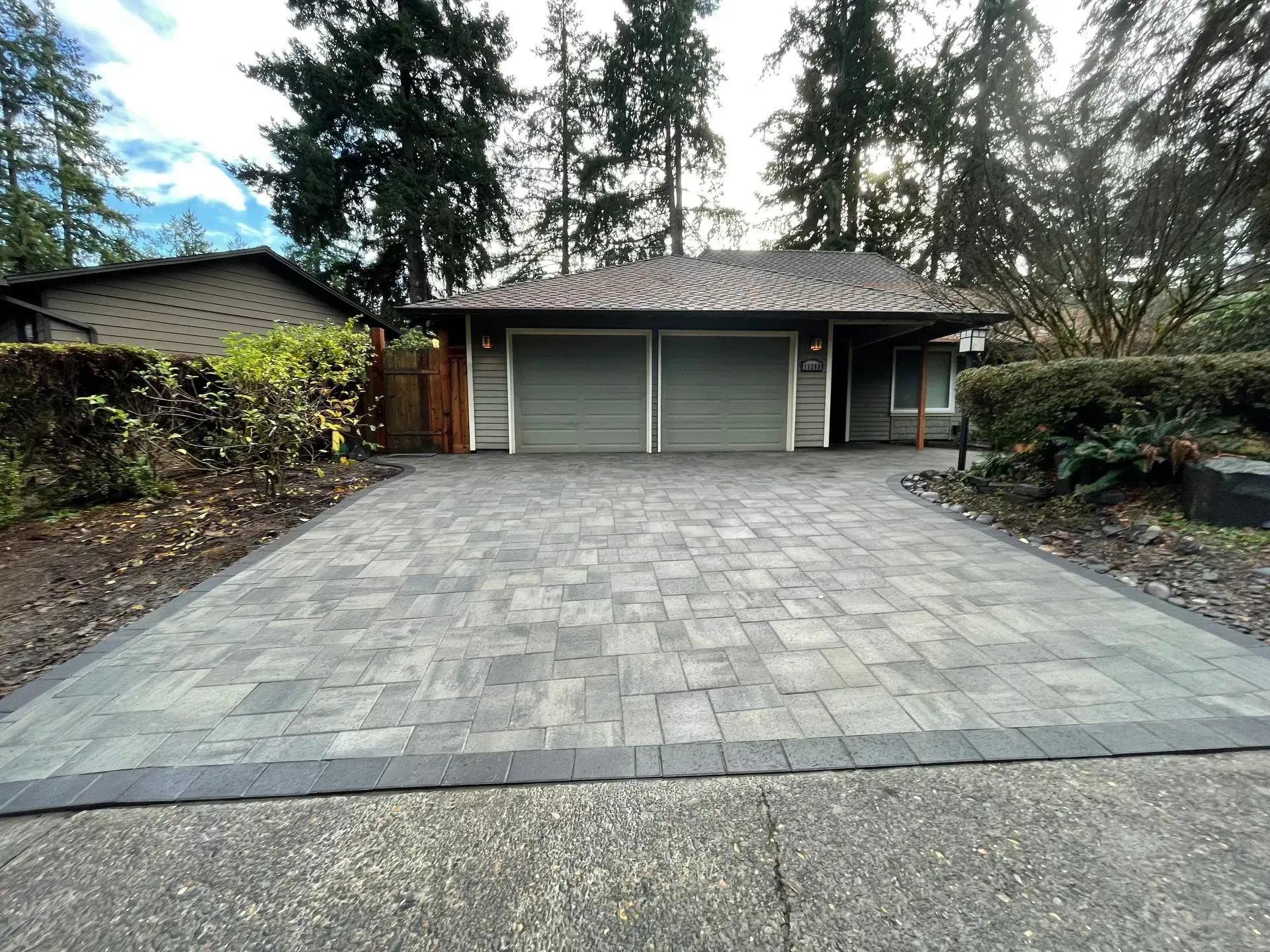 Gray paver driveway leads to a gray-painted house with two garage doors; evergreen trees in the background.