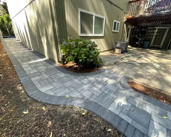 Paver walkway curves around a house, with landscaping and building in background.