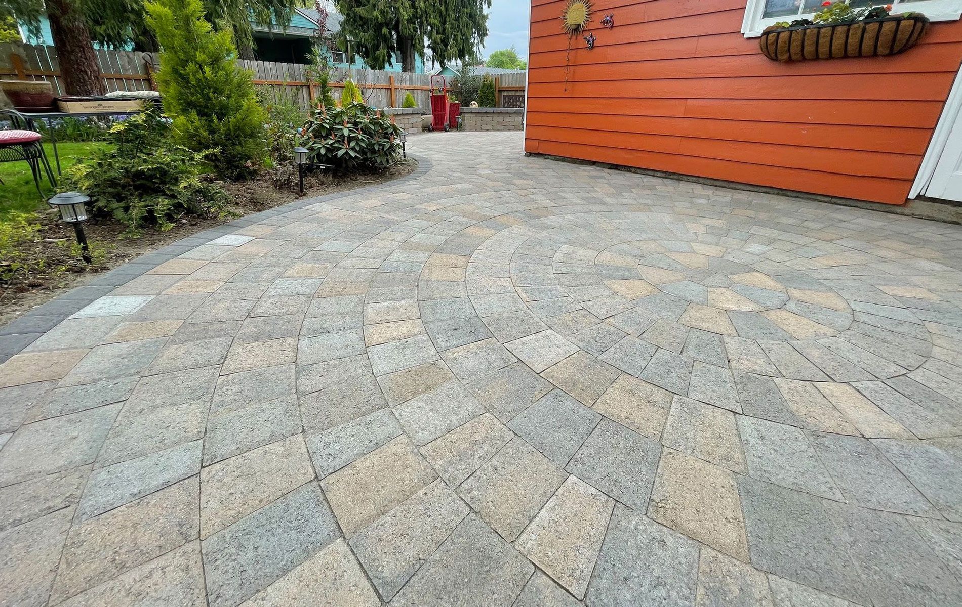 Stone patio with circular design next to orange house and garden.