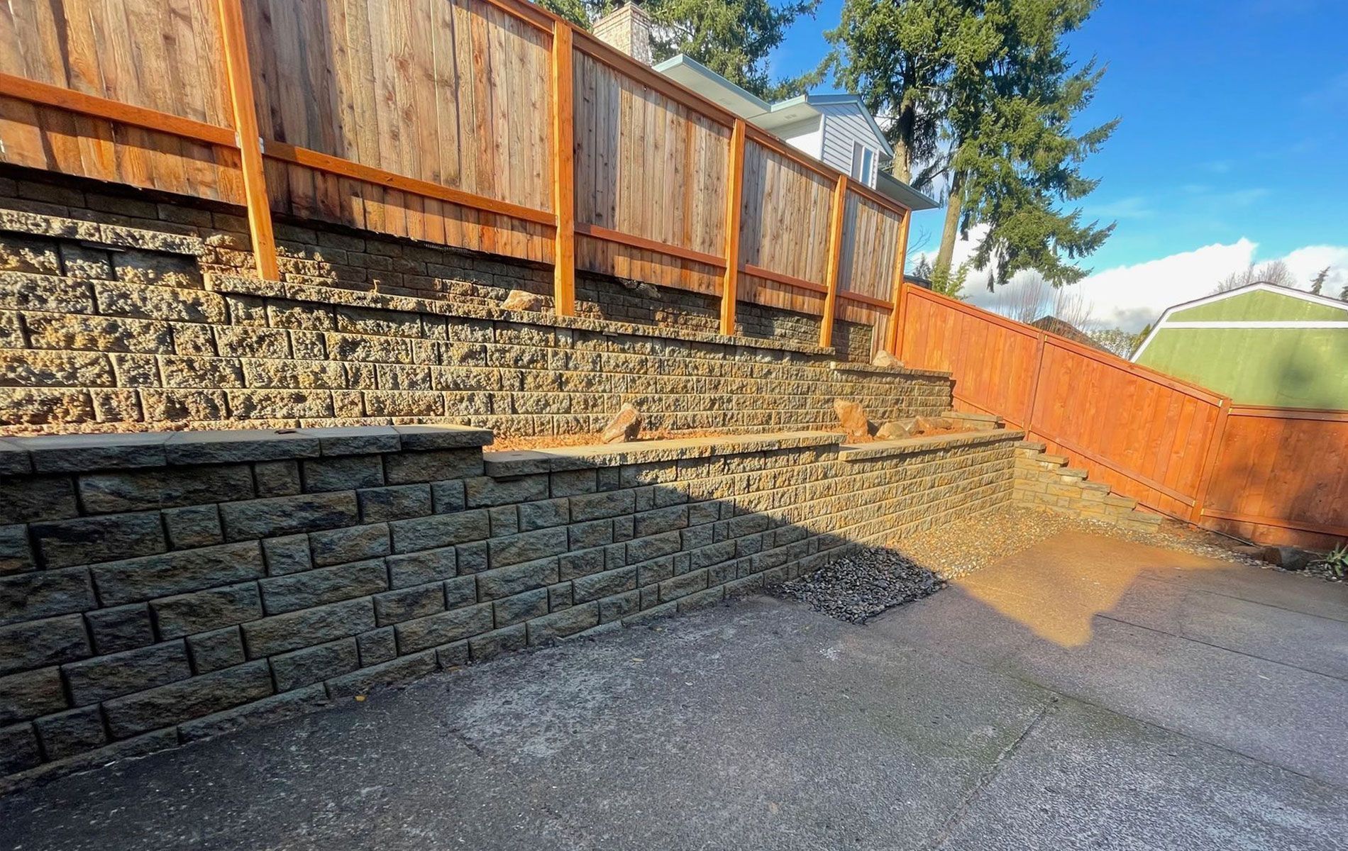 Retaining wall made of gray blocks supporting a wooden fence. Pathway slopes down from the fence.
