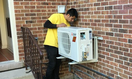 A man in a yellow shirt is checking an air conditioner HVAC on a brick wall.