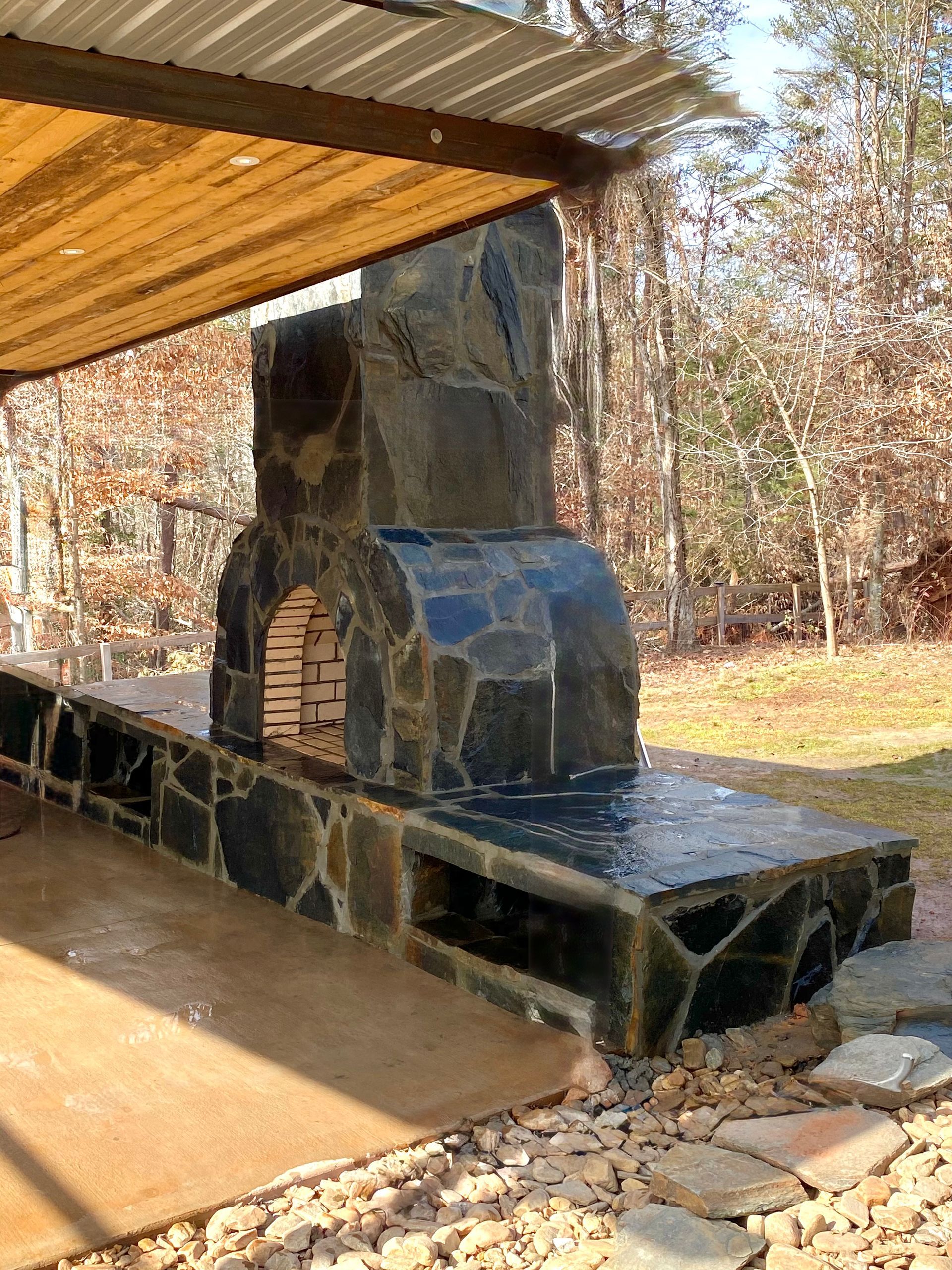 Outdoor stone oven and counter with a roof, set in a yard with trees.