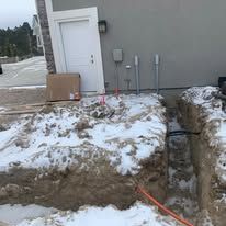 A trench with snow, and electrical conduits near a building door.