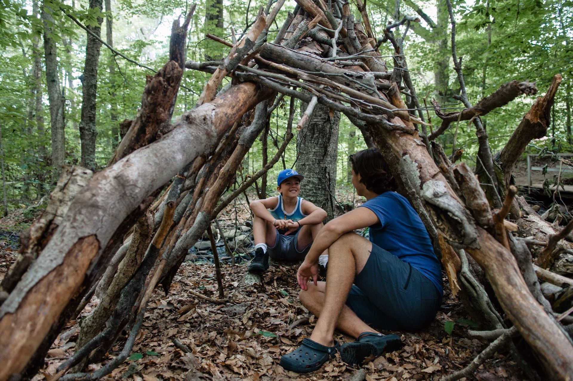 Two people are sitting in a hut made of branches in the woods.