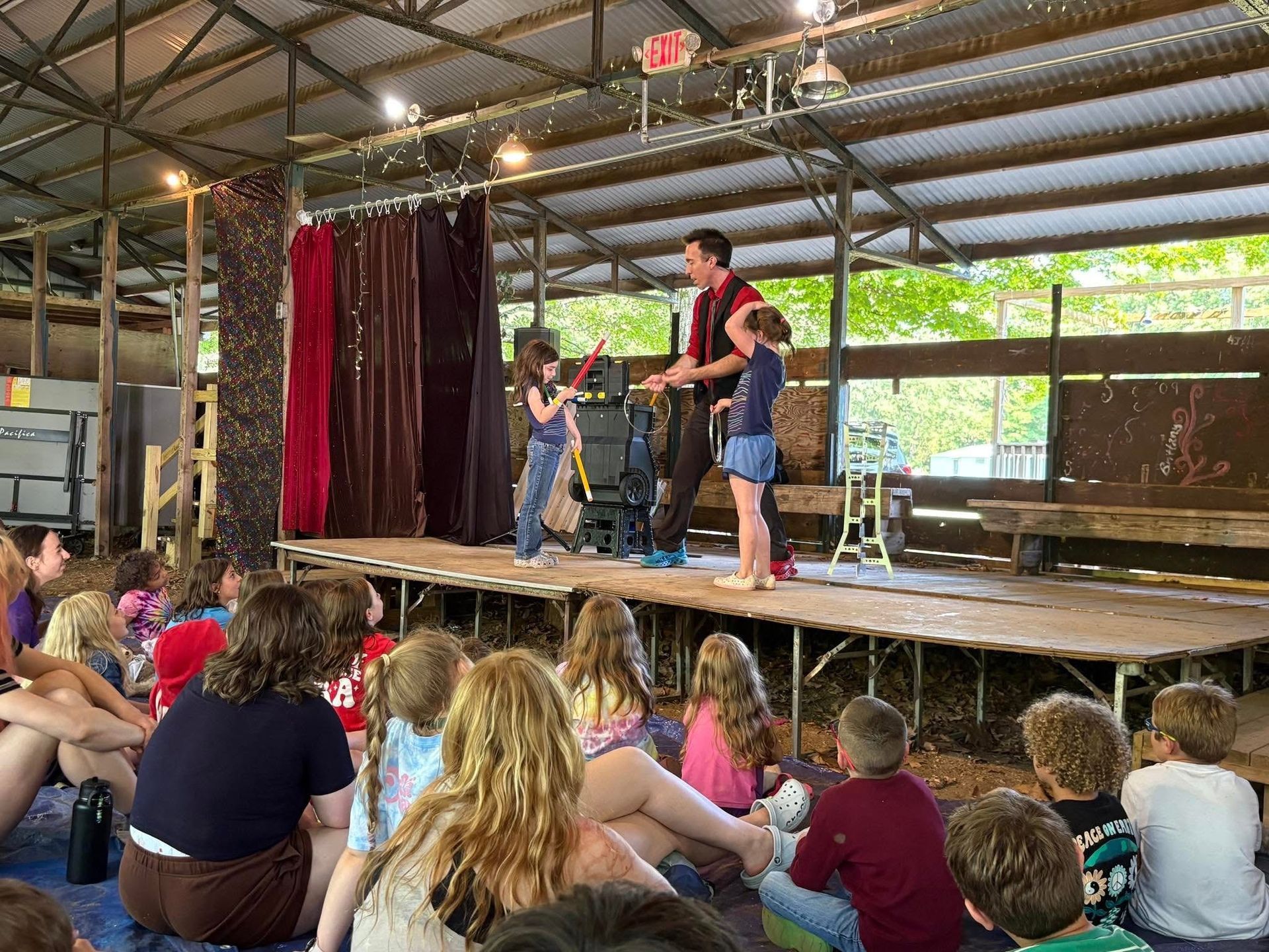 A group of children are sitting on the ground watching a man perform on a stage.