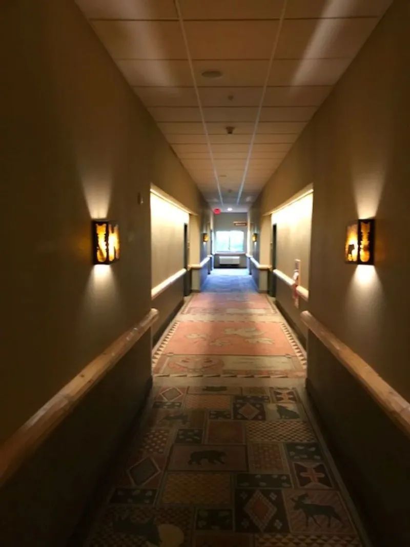 Hotel hallway with sconce lighting, long patterned carpet, doors on either side, and an exit at the end.