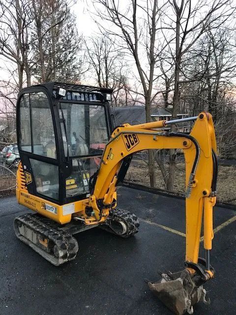 Yellow JCB mini excavator on a paved surface, arm extended with a bucket, in front of trees.
