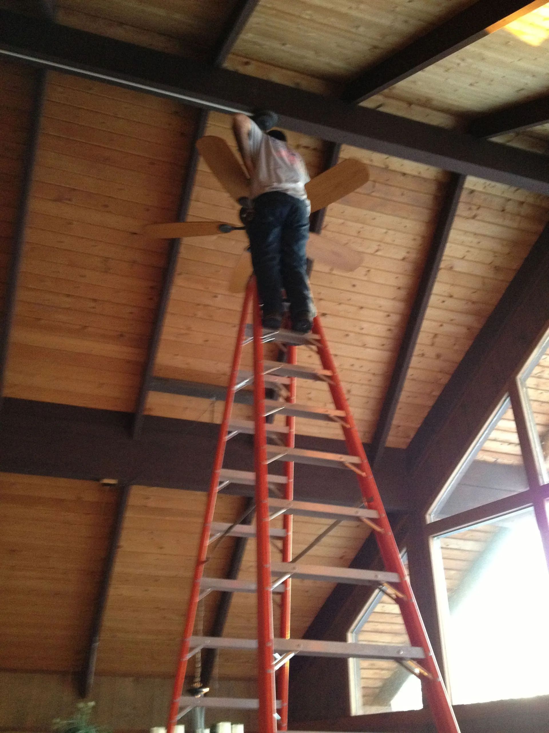 Person on tall orange ladder, installing ceiling fan on wood paneled ceiling.