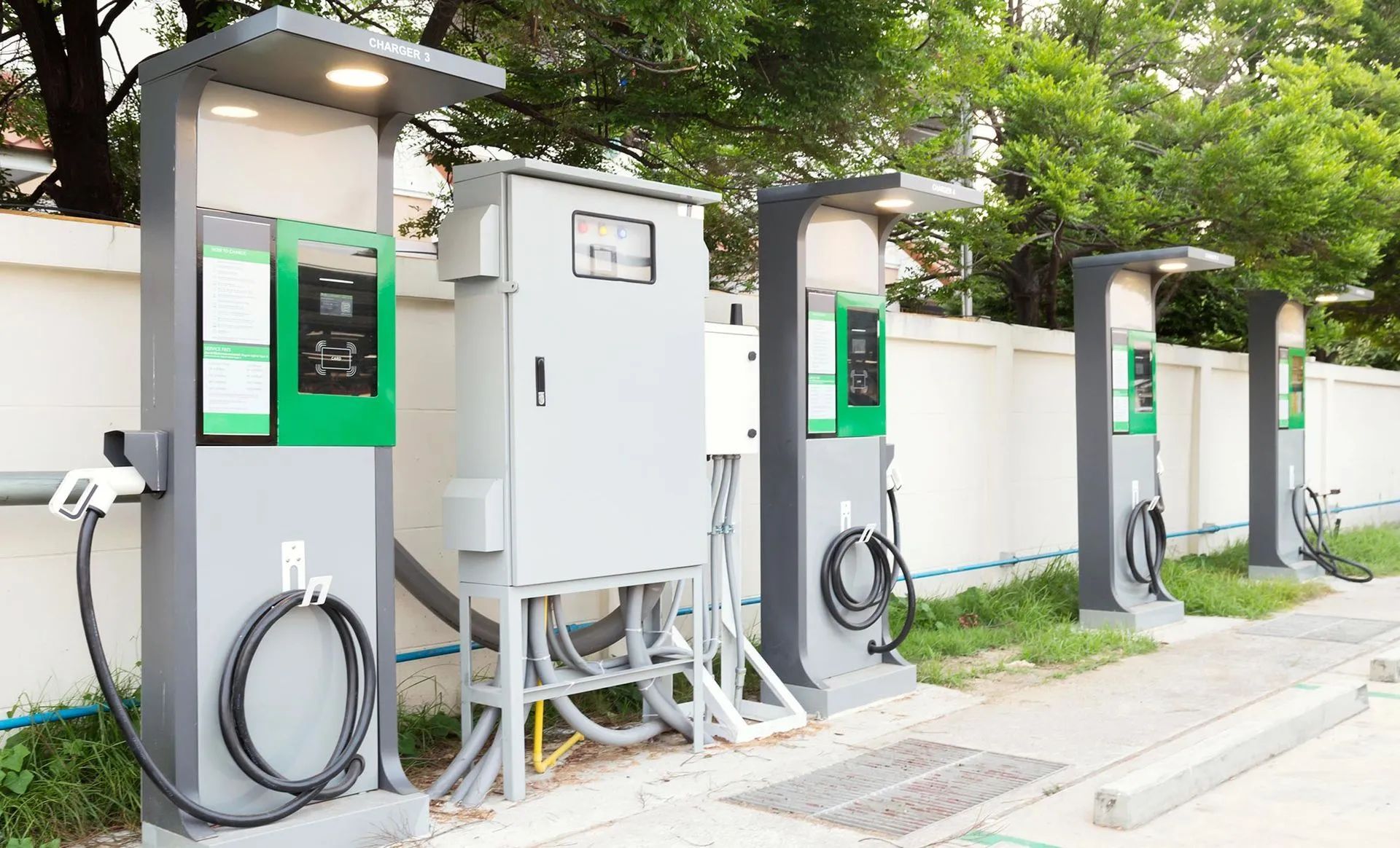 Electric vehicle charging stations along a white wall, gray and green color scheme, power cords attached.