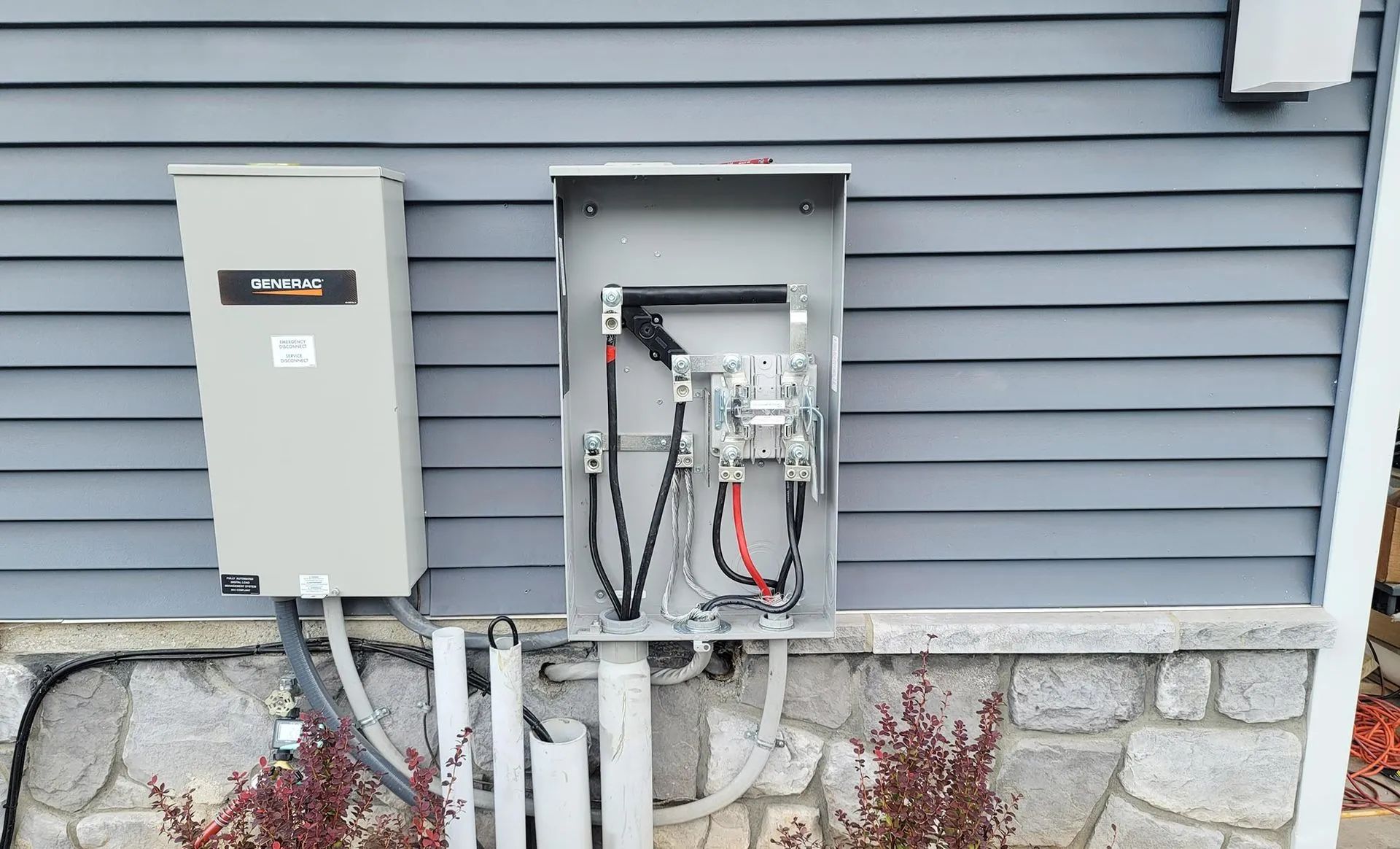 Gray electrical panel and meter box mounted on a light blue siding wall, over a stone foundation.