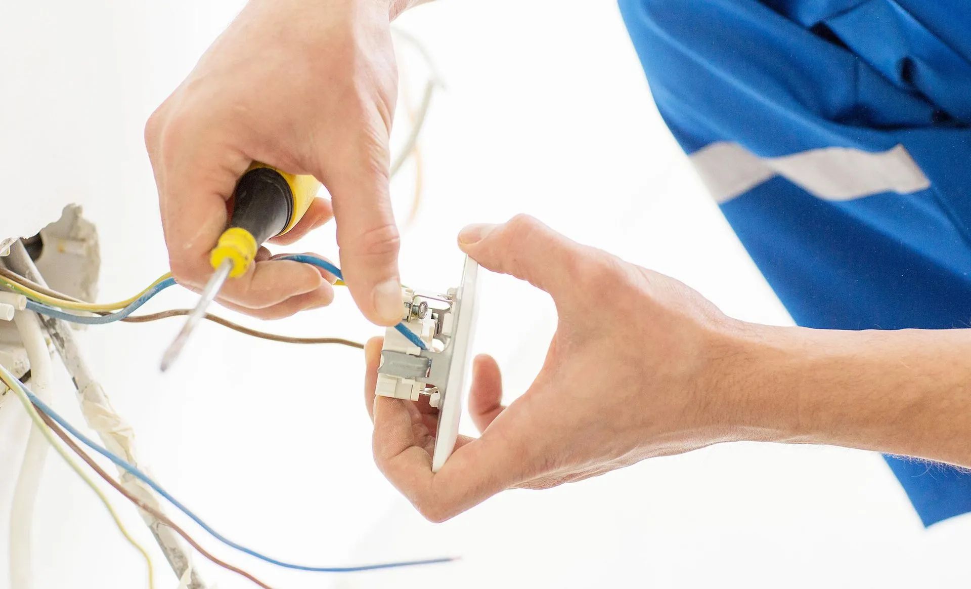 Electrician installing a white electrical outlet with a screwdriver, blue uniform, indoor setting.