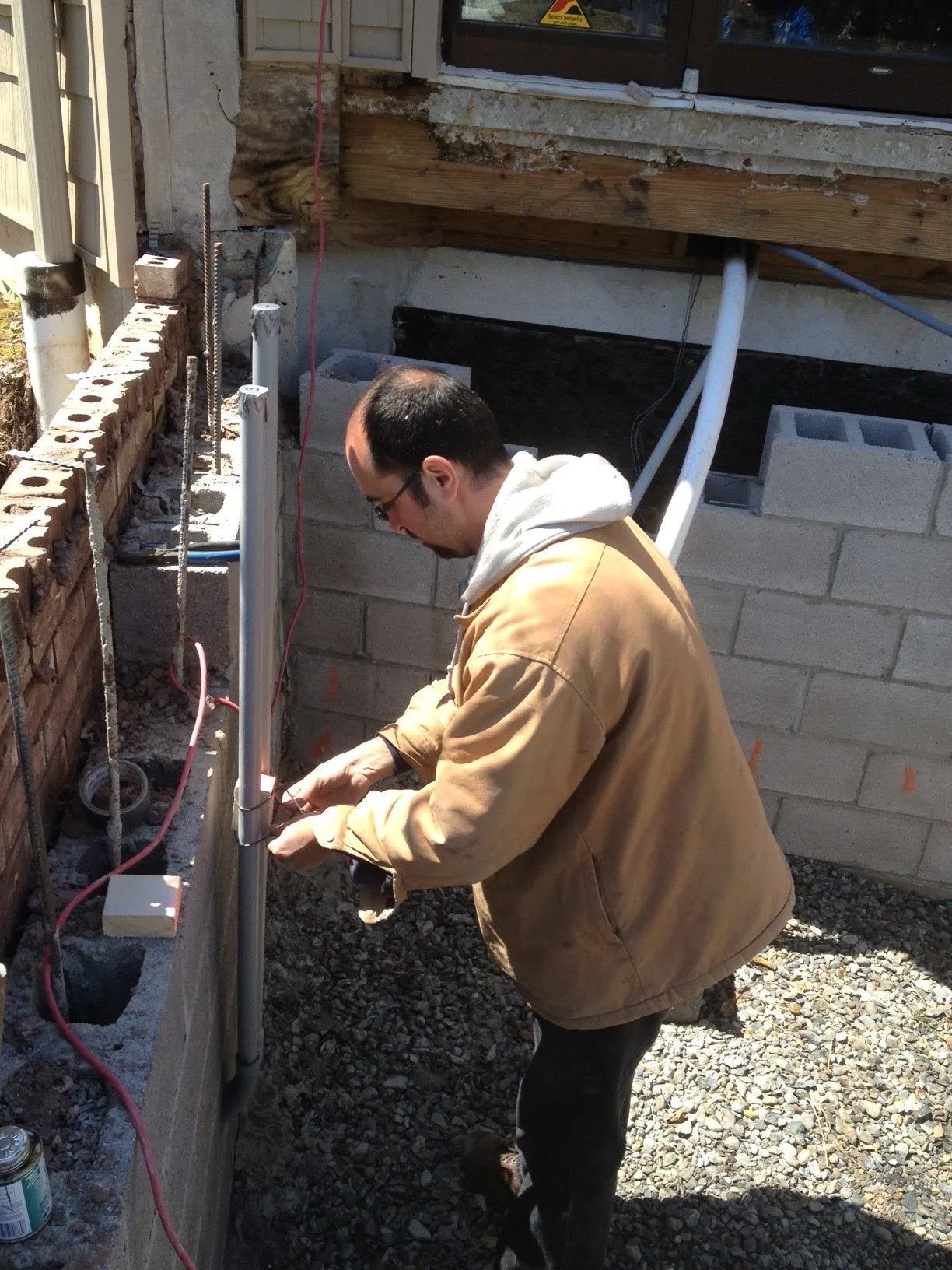 A person in a brown jacket working on a construction project, likely installing a metal post next to a concrete block wall.