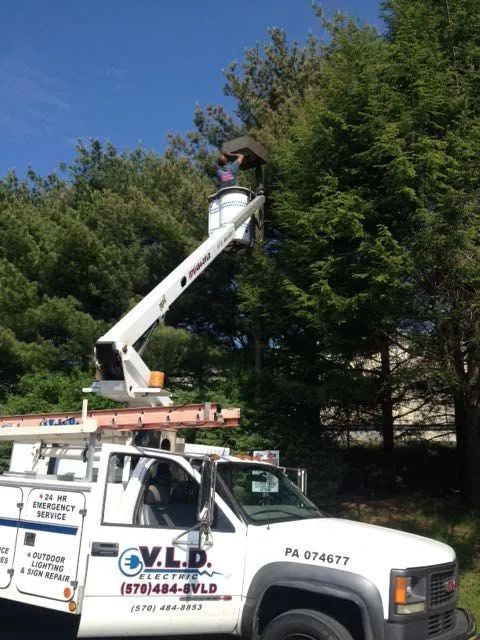 A worker in a bucket truck trims a tree. V.L.D. Electric truck is visible with PA license plate.