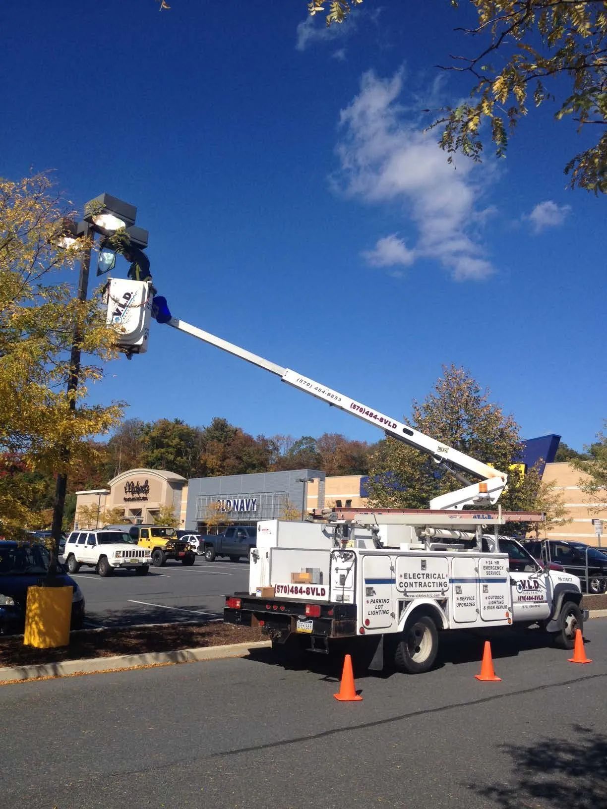 A utility truck with boom lift repairs a streetlight in a parking lot on a sunny day.