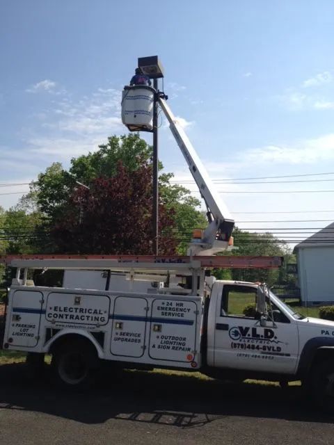 A white electrical contractor truck with a boom lift extending towards a light pole, against a blue sky.