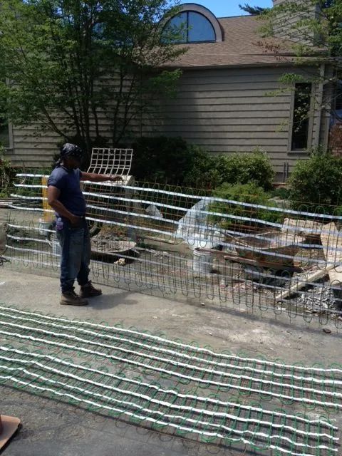 Man standing near a metal fence under construction in front of a house. Green rolls of material are on the ground.