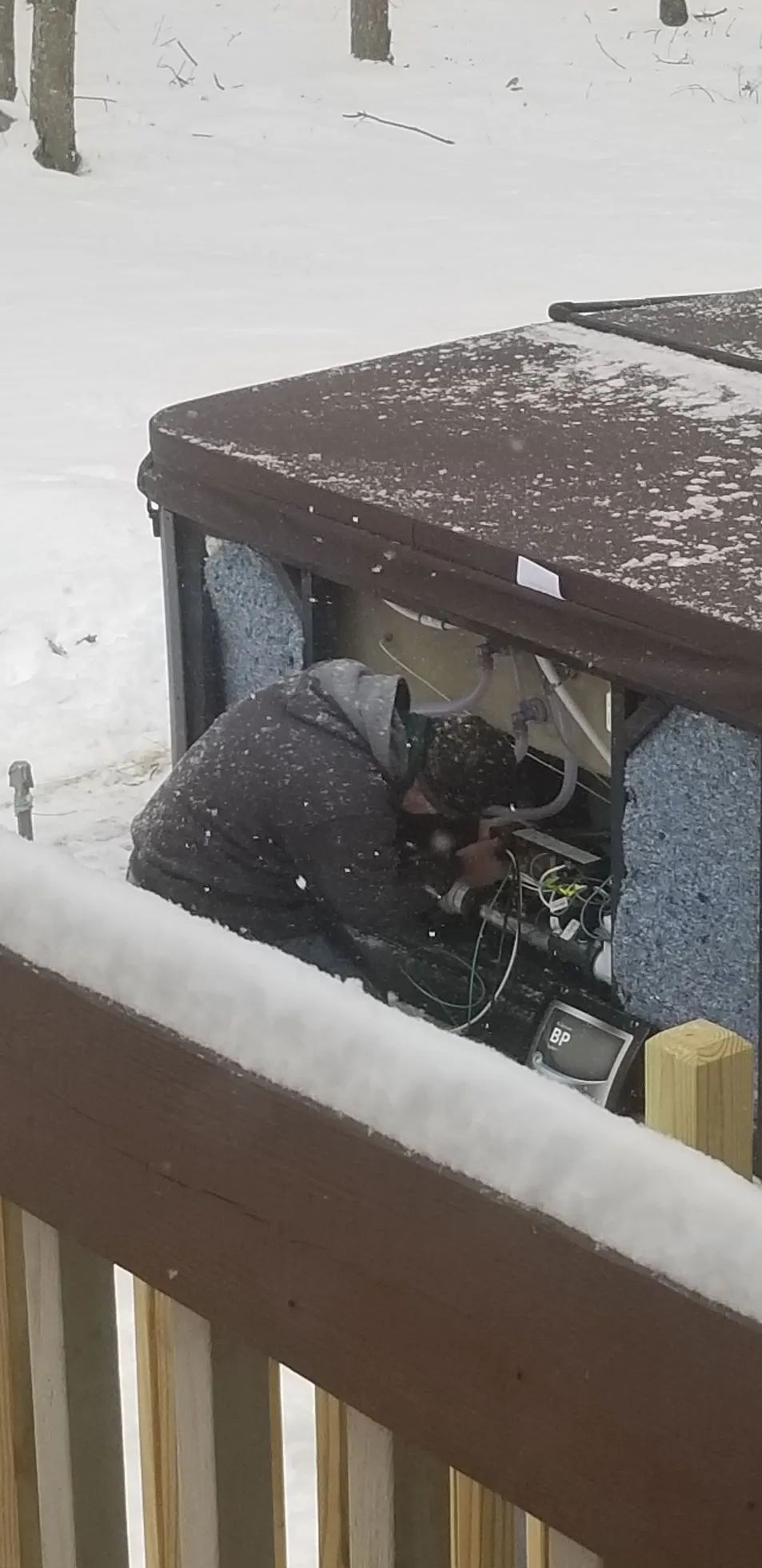 Person working on the interior of a hot tub on a snowy day. Brown deck railing.