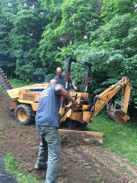 Two people operating a yellow backhoe on a dirt path next to a green wooded area.