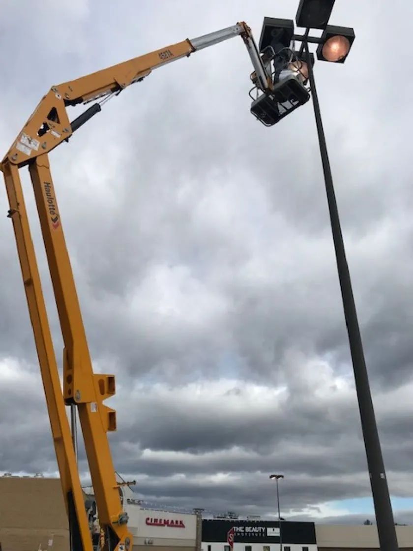 A person in a lift bucket repairs a tall streetlight. Yellow lift extends towards a dark pole under a cloudy sky.