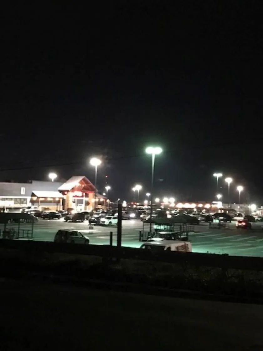 Night view of a brightly lit parking lot with a building in the background. Cars are parked throughout.