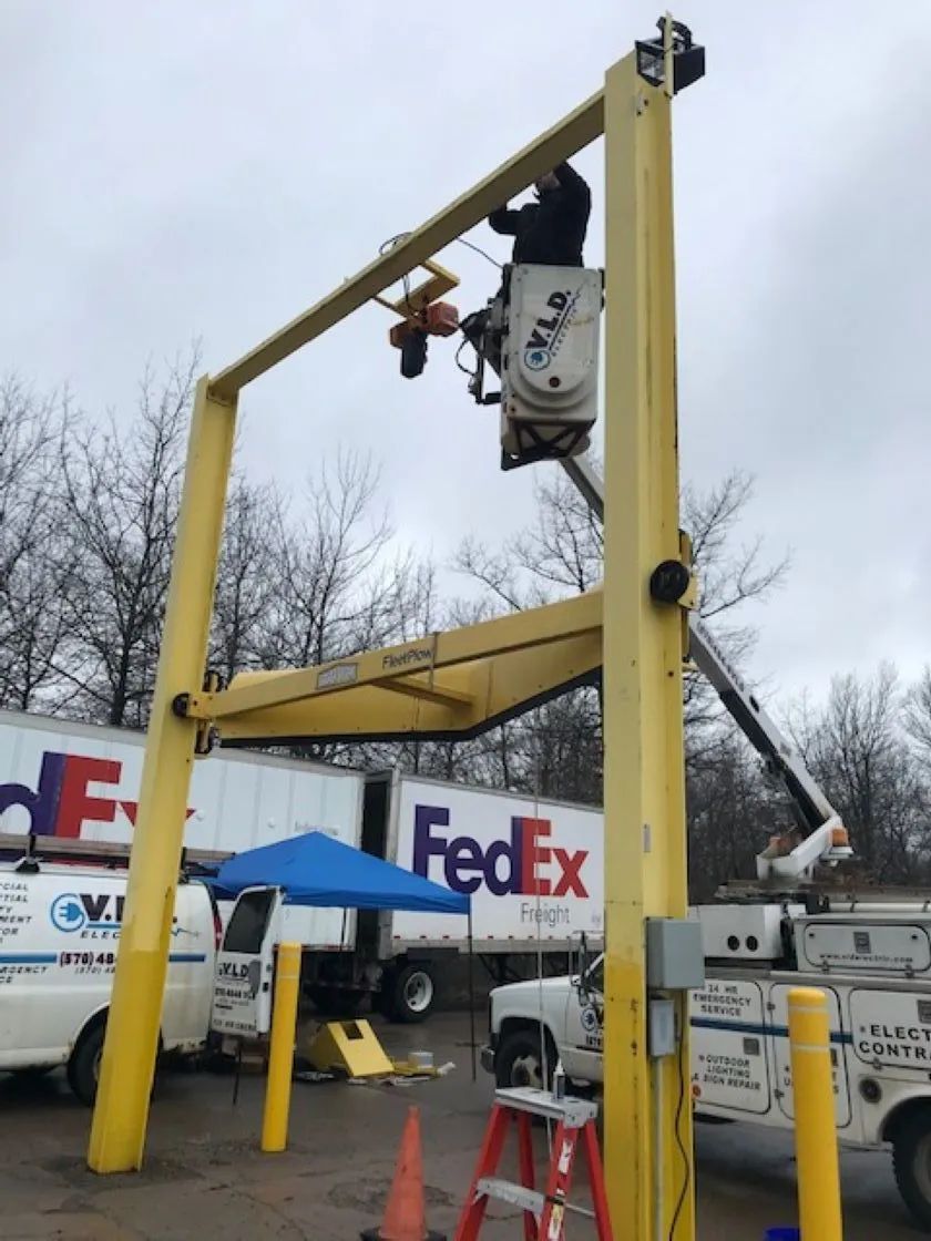 Person in aerial lift working on yellow overhead structure, FedEx trucks in background.
