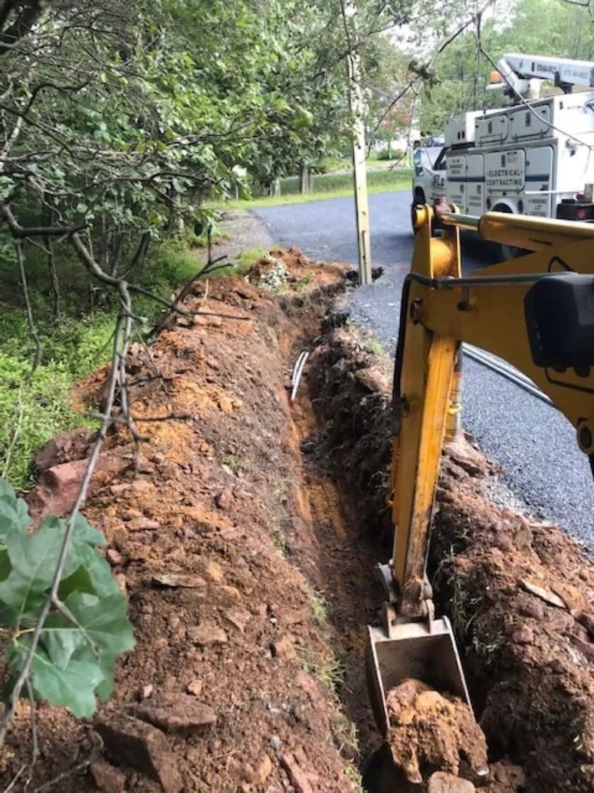 Yellow excavator digging a trench along a driveway. Dirt piles beside the trench.