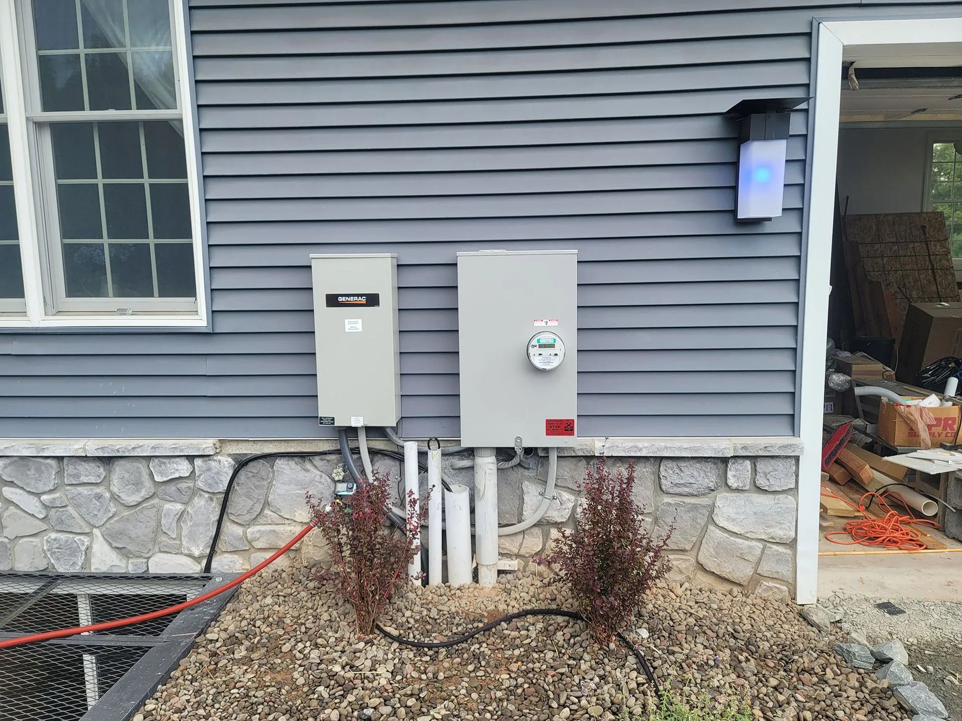 Electrical panel and meter on a gray house exterior, with a window and a blue light fixture.