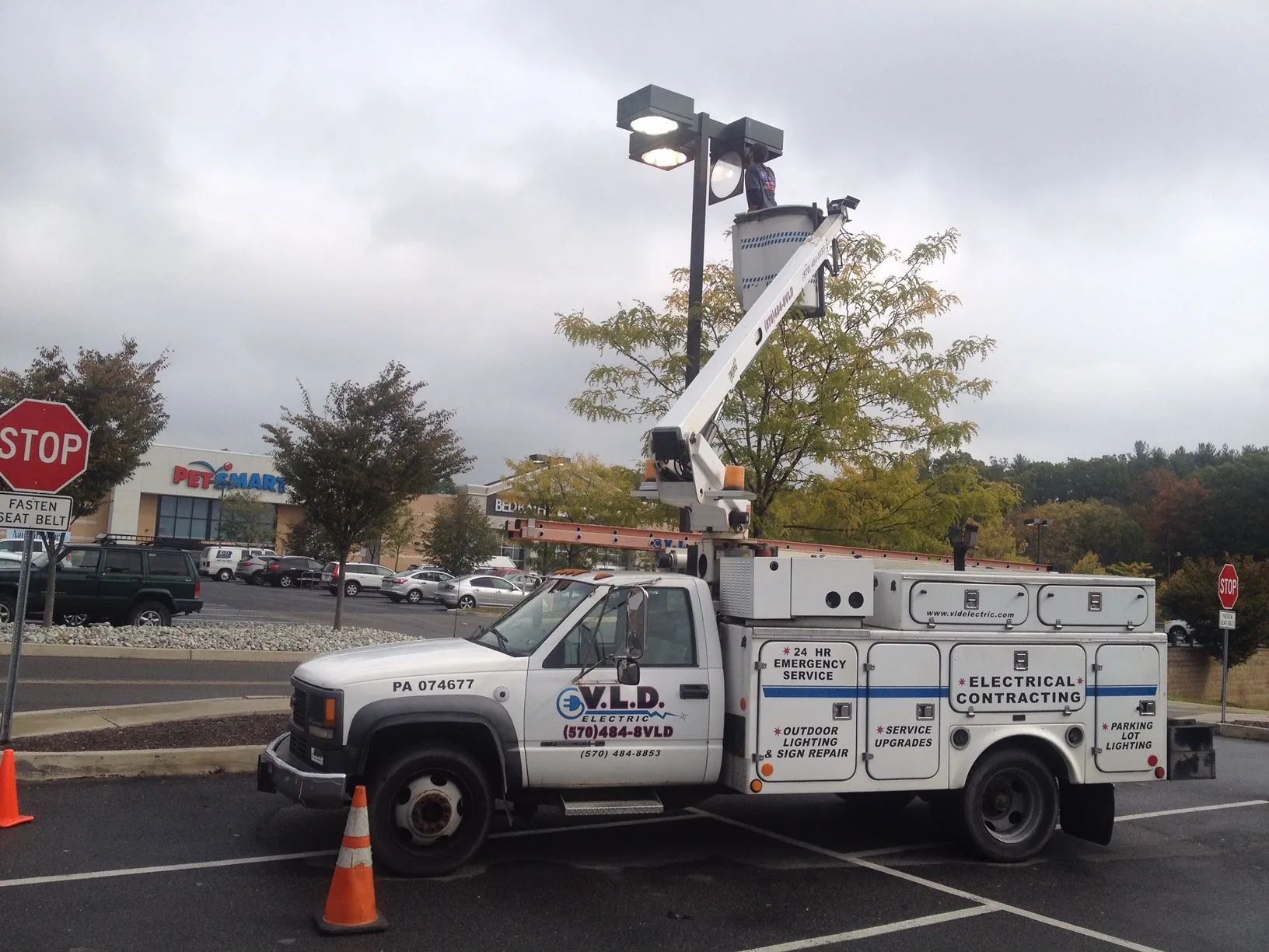 A utility truck with a lift arm extends toward a parking lot light post under a cloudy sky.
