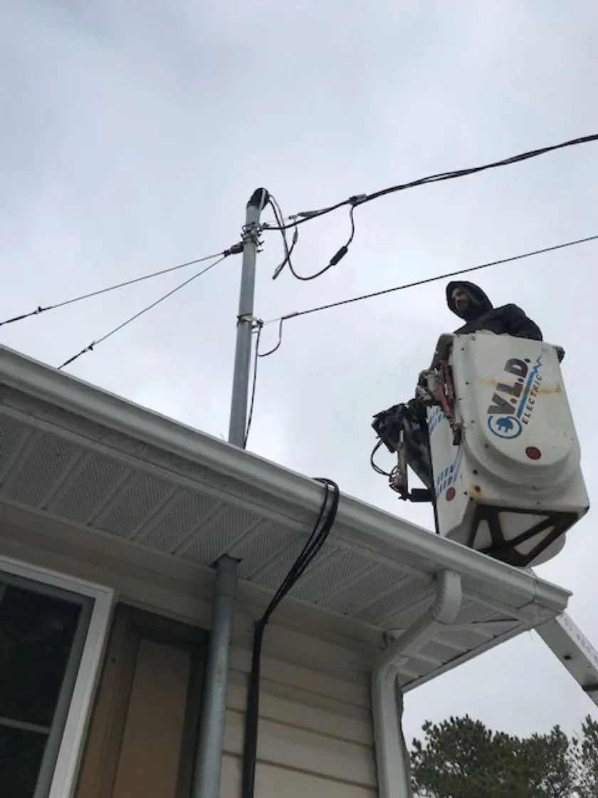 Two workers in a bucket truck near power lines. One works on a pole, against a cloudy sky.