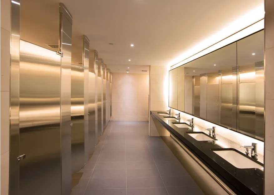 Modern public restroom with stainless steel stalls, sinks, large mirrors, and neutral-toned tiles.