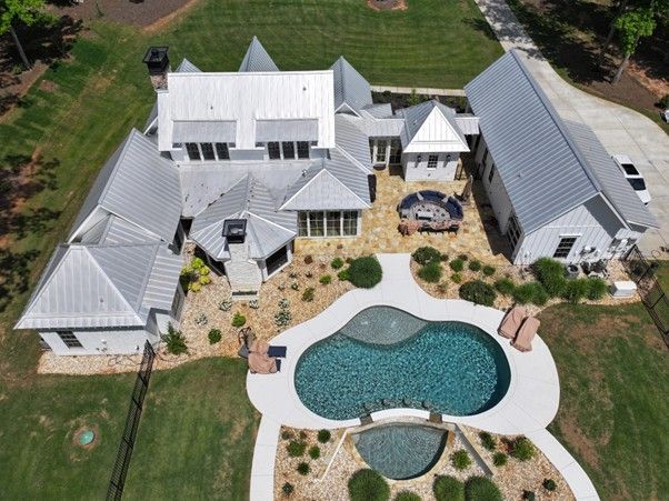 Aerial view of a white, multi-roof house with a pool, hot tub, stone patio, and green lawn on a sunny day.