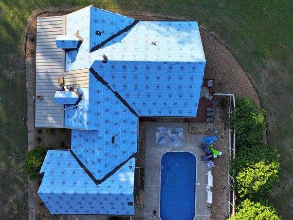 A top-down aerial view of a house undergoing roof repairs, featuring light blue underlayment and a nearby swimming pool.